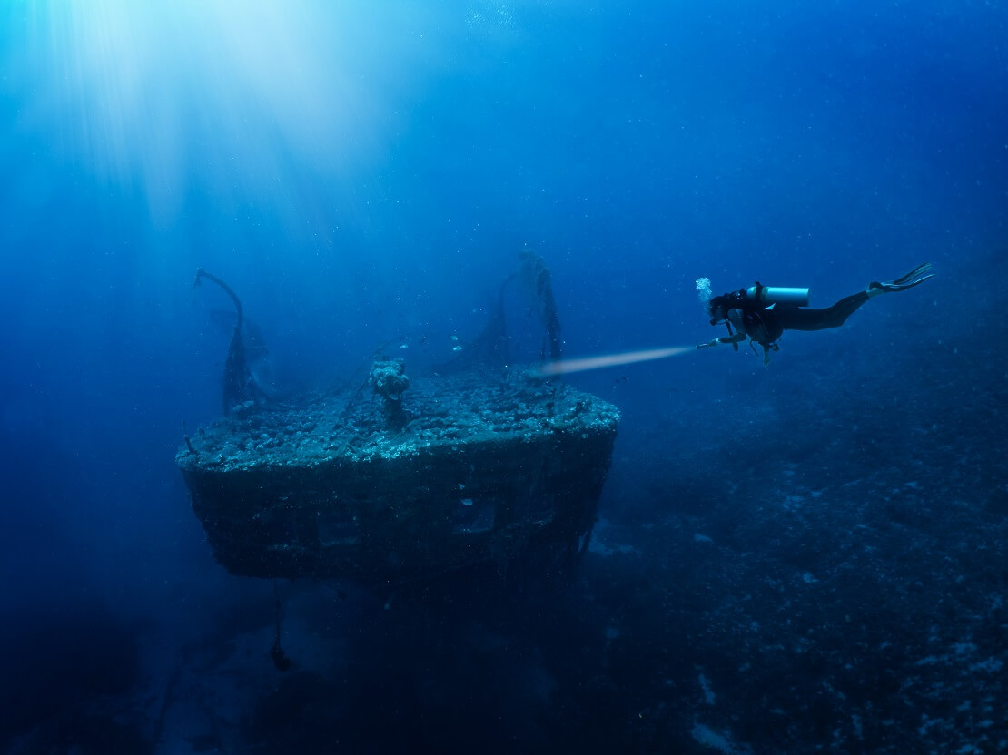 Taucher schwimmt mit einer Taschenlampe auf ein Schiffswrack zu