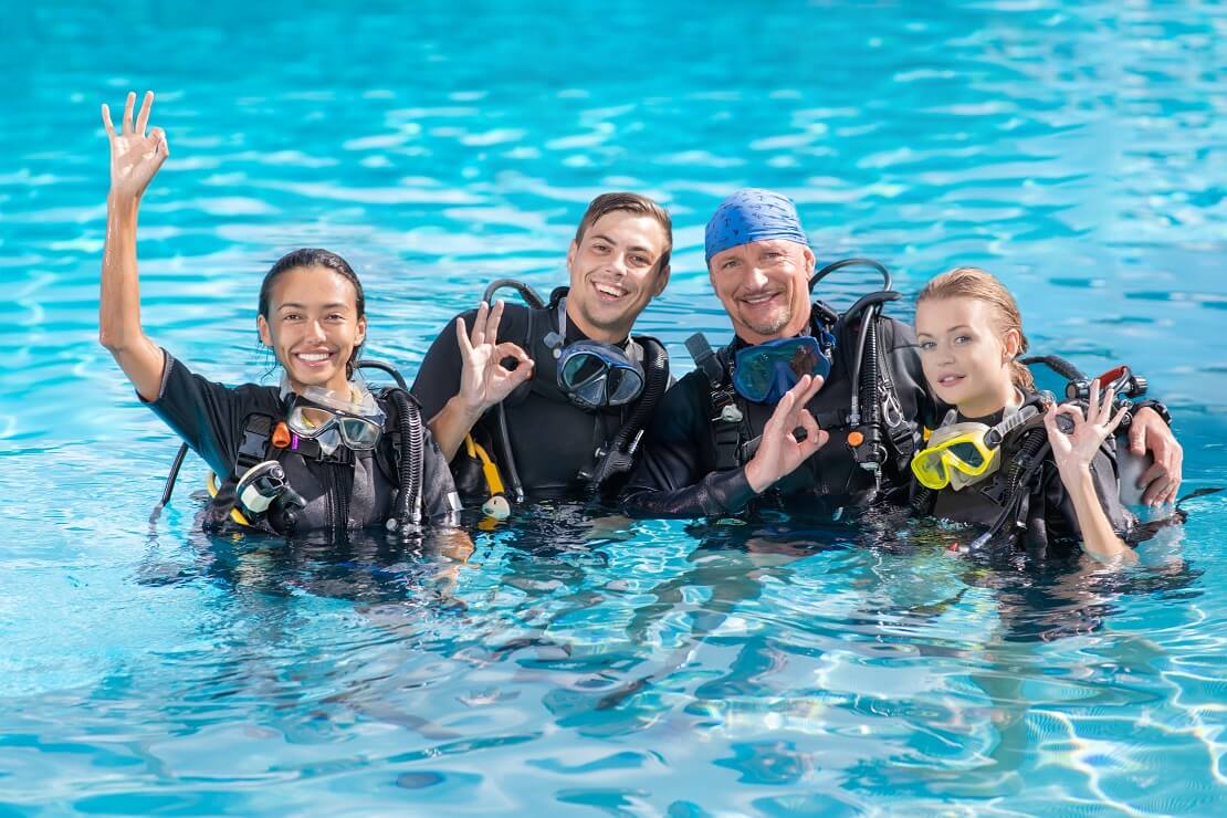 Vier Personen stehen zur hälfte im Wasser mit Tauchausrüstung und zeigen das alles ok ist