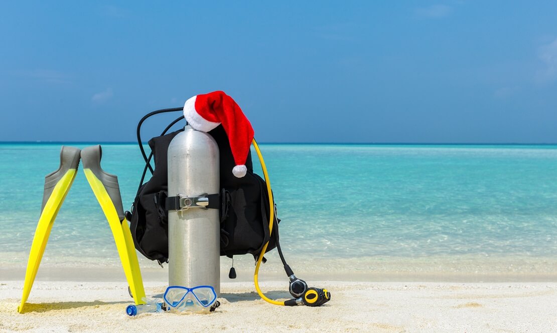 ABC Tauchausrüstung steht am Strand vor klarem Wasser auf der Pressluftflasche liegt eine Weihnachtsmütze